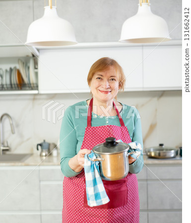 Elderly housewife in apron stands in kitchen in front of table and holds stainless pan with lunch 131662412