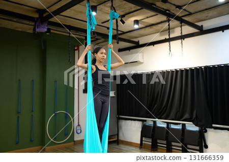 Woman practicing aerial yoga in fitness studio, standing inside blue hammock fabric for balance and strength training 131666359