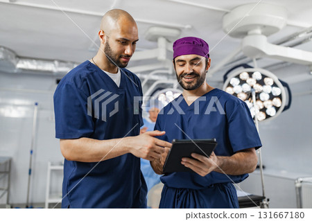 Two surgeons in an operating room review medical records on a tablet, discussing patient care. Two surgeons in an operating room review medical records on a tablet, discussing patient care. 131667180