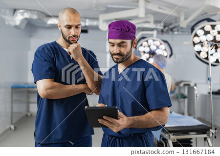 Two surgeons in an operating room review a tablet, likely discussing a patient's medical records before a procedure. Two surgeons in an operating room review a tablet, likely discussing a patient's medical records before a procedure. 131667184