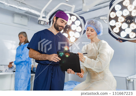 A surgeon reviews paperwork with a patient in an operating room, with another medical professional in the background. A surgeon reviews paperwork with a patient in an operating room, with another medical professional in the background. 131667223