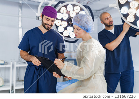 A surgeon reviews paperwork with a patient in an operating room, while another adjusts the surgical lights. A surgeon reviews paperwork with a patient in an operating room, while another adjusts the surgical lights. 131667226