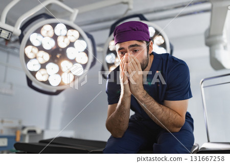 A stressed surgeon sits in an operating room, covering his face with his hands under bright surgical lights. A stressed surgeon sits in an operating room, covering his face with his hands under bright surgical lights. 131667258