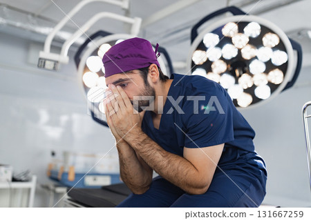 A stressed surgeon sits in an operating room, covering his face with his hands, illuminated by surgical lights. 131667259