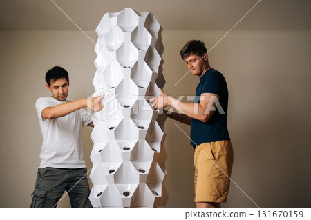 Front view of two men placing main body of vertical hydroponic tower garden onto its base, working together to set up indoor space for cultivating fresh plants. Concept of urban farming. Front view of two men placing main body of vertical hydroponic tower garden onto its base, working together to set up indoor space for cultivating fresh plants. Concept of urban farming. 131670159