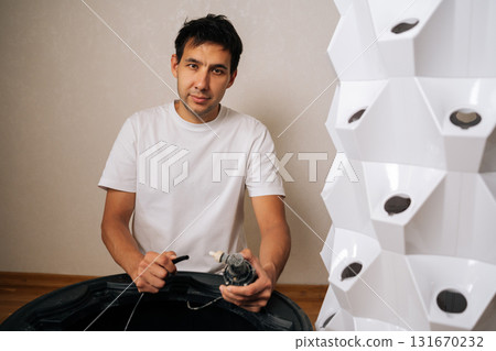 Portrait of home gardener connecting water pump to vertical hydroponic tower garden, setting up soilless indoor growing system for cultivating plants. Concept of healthy and sustainable cultivation. 131670232