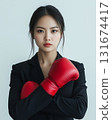 Confident young asian woman wearing red boxing gloves stands with arms crossed in a stylish black suit, displaying strength and determination 131674417