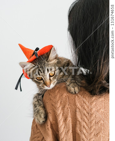 Woman in brown sweater holds tabby kitten wearing small witch's hat on her shoulder. Sweet festive atmosphere of Halloween highlights the bond between humans and animals Woman in brown sweater holds tabby kitten wearing small witch's hat on her shoulder. Sweet festive atmosphere of Halloween highlights the bond between humans and animals 131674658