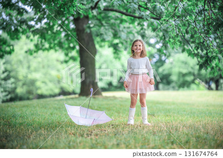 Little girl standing beside a closed umbrella and smiling gently at the camera. A calm and open moment of childhood presence in a rainy park. 131674764