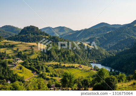 Lush green valley with rural houses surrounded by forested mountains under clear blue sky. 131675064