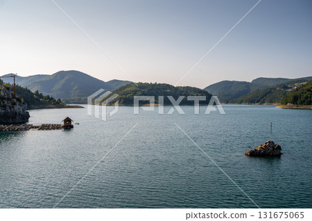 Rocky islet with a cross in peaceful blue lake, surrounded by green forest hills under clear sky 131675065