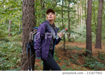 Young female traveler leaning on tree with water bottle and backpack during hike 131677901
