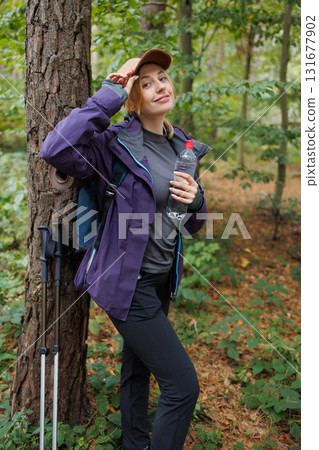 Female hiker adjusting cap while holding water bottle and smiling in forest 131677902