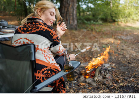 Camper woman resting in chair with drink near forest campfire Camper woman resting in chair with drink near forest campfire 131677911
