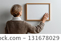 An artwork mockup being hung by a young woman. Rear view of a girl with a large poster frame on the wall, an empty blank frame in a modern apartment. 131678029