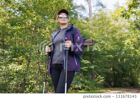 Confident female hiker with trekking poles walking on forest path Confident female hiker with trekking poles walking on forest path 131678143