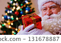 portrait of a happy and Jolly Santa Claus holding a gift in front of a Christmas tree. Christmas spirit, holidays and celebrations concept. 131678887