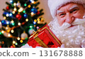 portrait of a happy and Jolly Santa Claus holding a gift in front of a Christmas tree. Christmas spirit, holidays and celebrations concept. 131678888