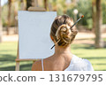 Young woman sitting outdoors with a paintbrush tucked into her hair bun, facing a blank canvas on an easel. Concept of creativity, inspiration, and artistic lifestyle. 131679795