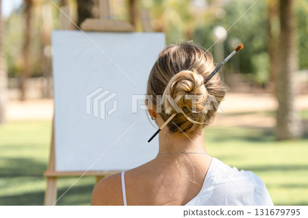 Young woman sitting outdoors with a paintbrush tucked into her hair bun, facing a blank canvas on an easel. Concept of creativity, inspiration, and artistic lifestyle. Young woman sitting outdoors with a paintbrush tucked into her hair bun, facing a blank canvas on an easel. Concept of creativity, inspiration, and artistic lifestyle. 131679795