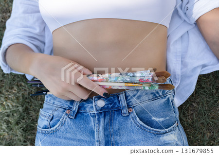 Closeup of a woman's hand tucking colourful used paintbrushes into blue jeans waistband, creative lifestyle detail with texture and casual fashion vibe, macro crop. Closeup of a woman's hand tucking colourful used paintbrushes into blue jeans waistband, creative lifestyle detail with texture and casual fashion vibe, macro crop. 131679855