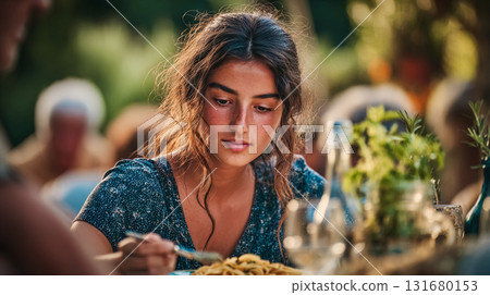 Young Italian woman enjoying a meal outdoors with friends and family. A beautiful girl with long curly hair at an outdoor dinner, looking down at plate. Family lunch. This is AI-generated item 131680153