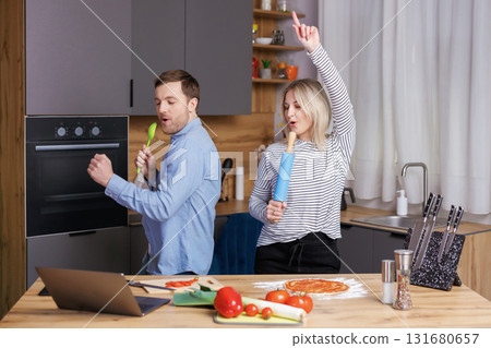 Home concert. Playful spouses having fun in kitchen, singing and dancing. Cheerful couple using kitchen utensils as microphones, fooling while cooking lunch at home. Home concert. Playful spouses having fun in kitchen, singing and dancing. Cheerful couple using kitchen utensils as microphones, fooling while cooking lunch at home. 131680657