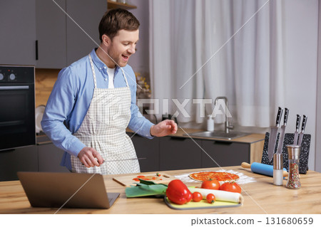 Young handsome man is preparing pizza and salad in the kitchen. The man enjoys the process of cooking. Young handsome man is preparing pizza and salad in the kitchen. The man enjoys the process of cooking. 131680659