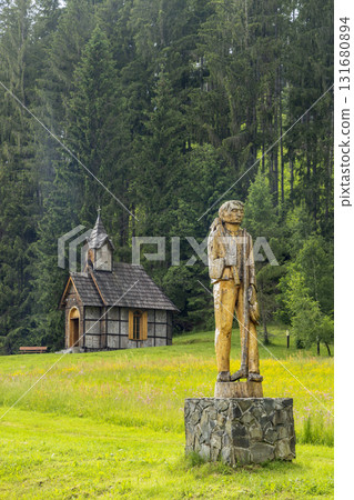 Wooden sculpture of man in meadow with rustic chapel and forest 131680894
