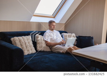 Man working on laptop and using smartphone while sitting on sofa at home. Concept of freelancing, digital lifestyle, mobile technology, e-banking, online shopping, communication and productivity.  131682425