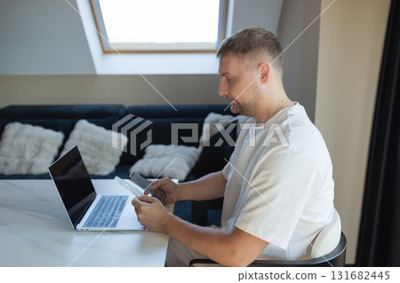 Man working remotely at home with laptop and smartphone, making online payment with credit card. Concept of digital banking, fintech, e-commerce, modern lifestyle and financial management. 131682445
