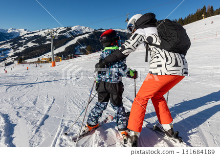 Mother teaching child to ski on sunny mountain slope, holding and guiding for balance and confidence. Warm family moment of encouragement and teamwork in bright winter landscape full of joy Mother teaching child to ski on sunny mountain slope, holding and guiding for balance and confidence. Warm family moment of encouragement and teamwork in bright winter landscape full of joy 131684189