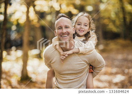 Mature father giving piggyback ride to small daughter on a walk in autumn forest. 131684624