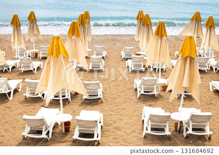 Beach scene with empty closed umbrellas and sun loungers on on sea shore Beach scene with empty closed umbrellas and sun loungers on on sea shore 131684692