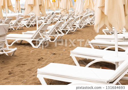 Beach scene with empty closed umbrellas and sun loungers on on sea shore Beach scene with empty closed umbrellas and sun loungers on on sea shore 131684693