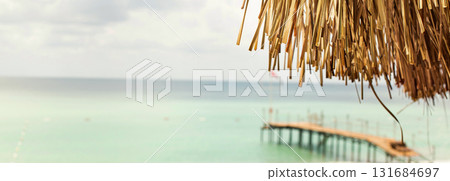Thatched roof with tropical beach with sand and wooden pier on the sea shore in background. 131684697