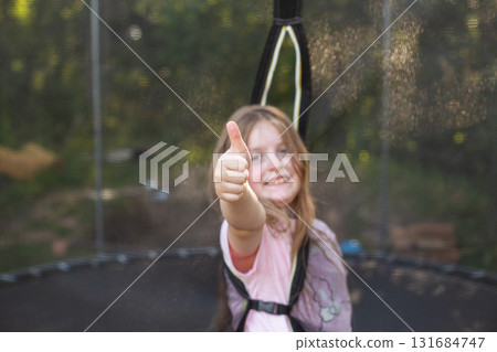 Happy excited child girl sitting on trampoline outdoor showing thumb up sign 131684747