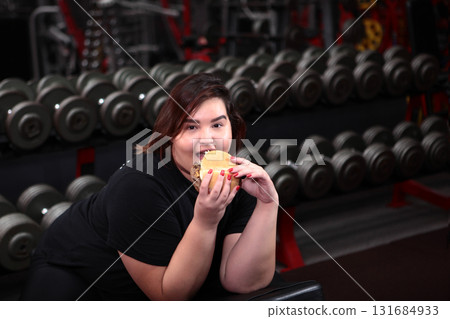 Fat, cheerful young woman of large size eats hamburger and does sports. Problem of being overweight. Concept of body positive. 131684933