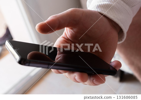 A man holds a smartphone in his hands. The phone has a sleek design and a dark screen. The background is blurred, focusing on the phone and hands. A man holds a smartphone in his hands. The phone has a sleek design and a dark screen. The background is blurred, focusing on the phone and hands. 131686085