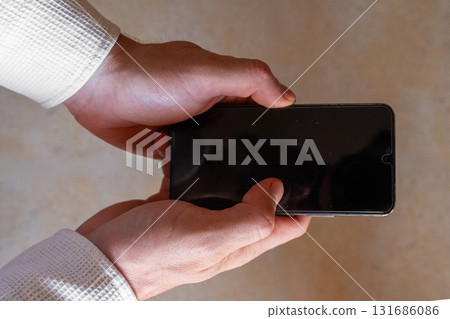 A man holds a smartphone in his hands. The phone has a sleek design and a dark screen. The background is blurred, focusing on the phone and hands. A man holds a smartphone in his hands. The phone has a sleek design and a dark screen. The background is blurred, focusing on the phone and hands. 131686086