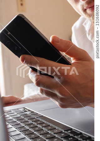 A man holds a smartphone in his hands. The phone has a sleek design and a dark screen. The background is blurred, focusing on the phone and hands. 131686092