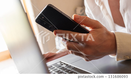 A man holds a smartphone in his hands. The phone has a sleek design and a dark screen. The background is blurred, focusing on the phone and hands. A man holds a smartphone in his hands. The phone has a sleek design and a dark screen. The background is blurred, focusing on the phone and hands. 131686093