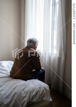 Older man sitting alone in emotional burden mental fatigue and deep depression by window, rearview Older man sitting alone in emotional burden mental fatigue and deep depression by window, rearview 131687170