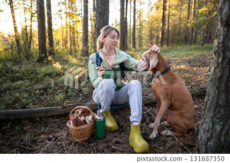 Happy serene woman petting calm dog while drinking tea during forest mushroom foraging walk 131687350