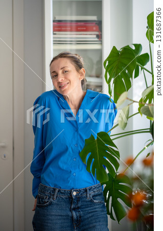Portrait of charming smiling middle aged woman stands next to monstera leaves at cozy home 131687366