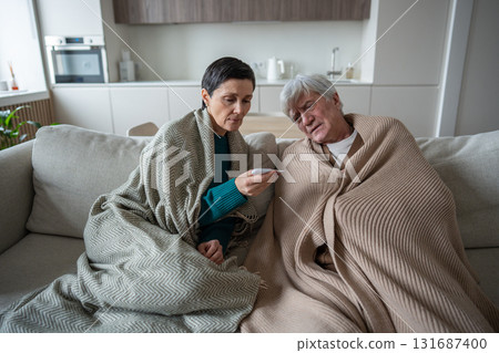 Concerned woman with thermometer in blanket check fever with struggling husband on couch at home 131687400
