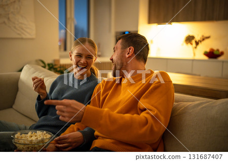 Smiling young couple enjoying popcorn watching movie together on living room sofa, family night Smiling young couple enjoying popcorn watching movie together on living room sofa, family night 131687407