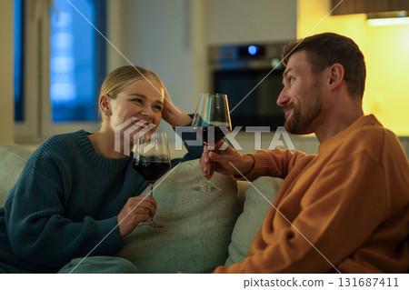 Happy couple enjoying wine together in cozy living room sharing intimate talk on couch at night Happy couple enjoying wine together in cozy living room sharing intimate talk on couch at night 131687411