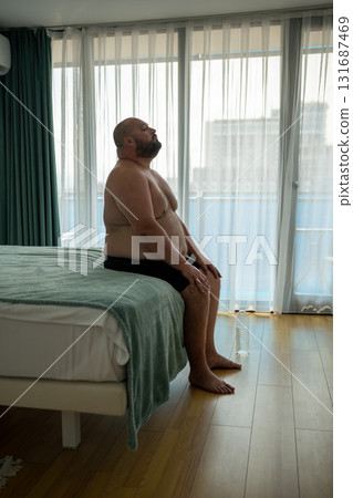 Overweight bare chested man sitting on bad with air conditioning on trying to cope with overheating 131687469