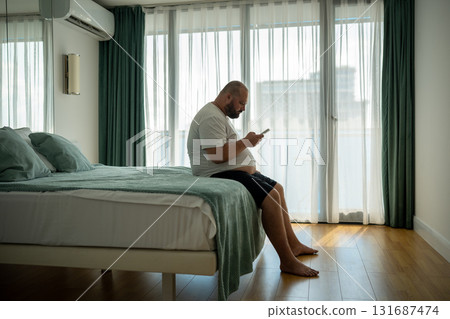 Overweight guy hanging in smartphone on resort sitting in bed in hotel room on vacation. 131687474